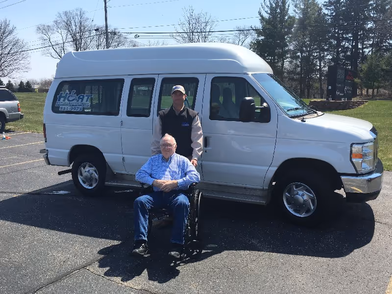 Two men, one seated in a wheelchair, pose beside a white transport van in parking lot.