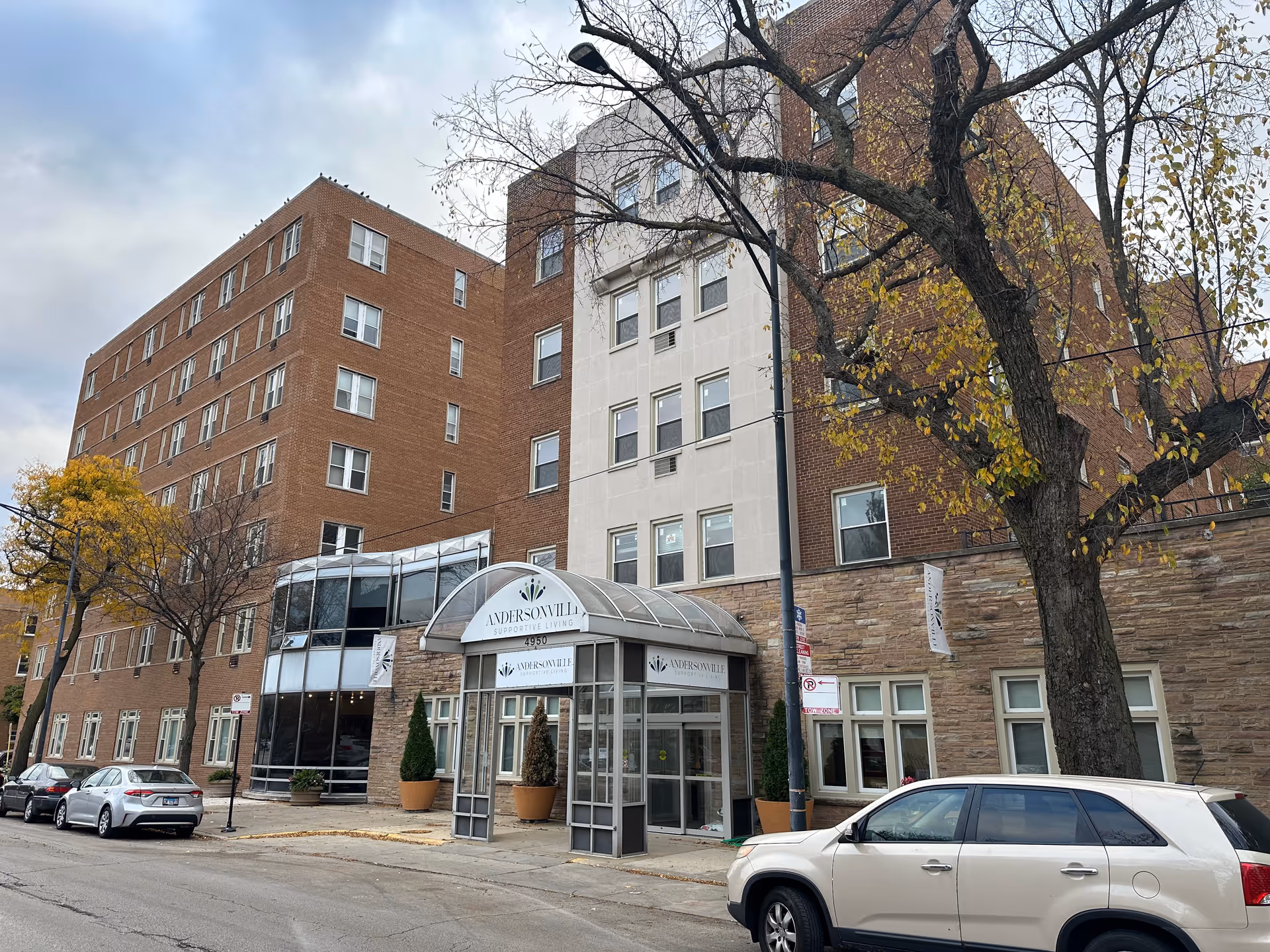 Exterior view of a multi-story brick and stone building with a glass entrance canopy labeled 'Andersonville Supportive Living'. Several cars are parked along the street in front of the building, and trees with autumn leaves are visible.