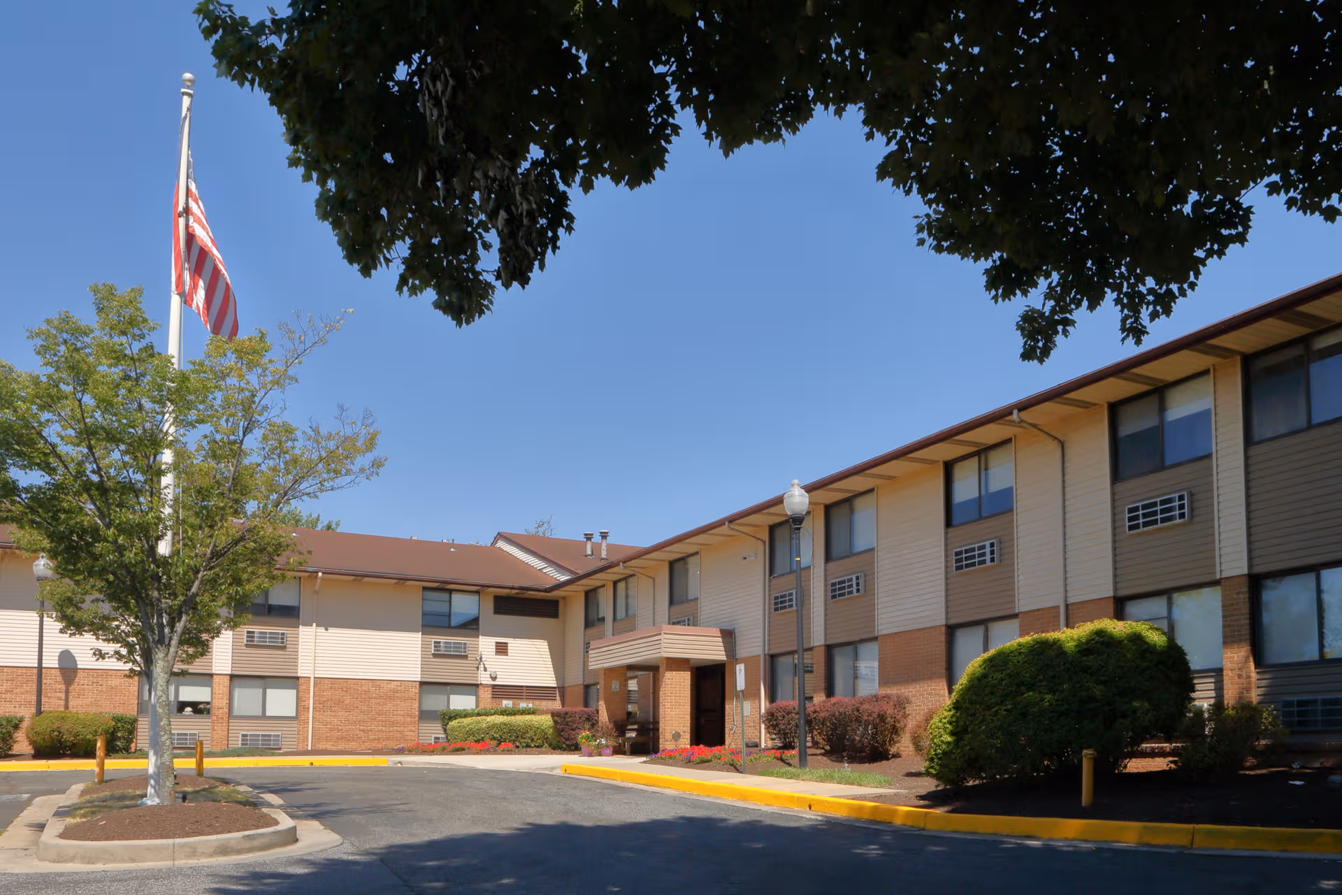 Two-story senior living facility exterior with flag, trees, and a parking lot.