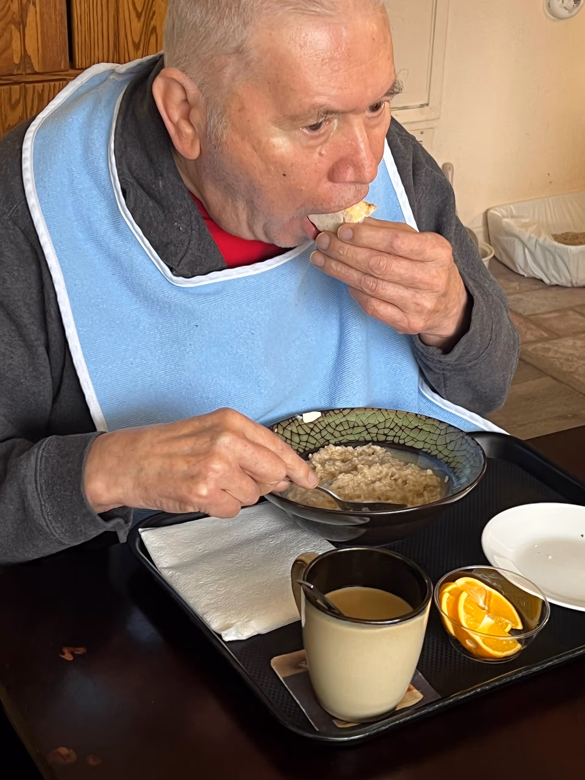 An older adult wearing a blue bib eating oatmeal with a piece of toast at a table with a tray holding coffee and orange slices.