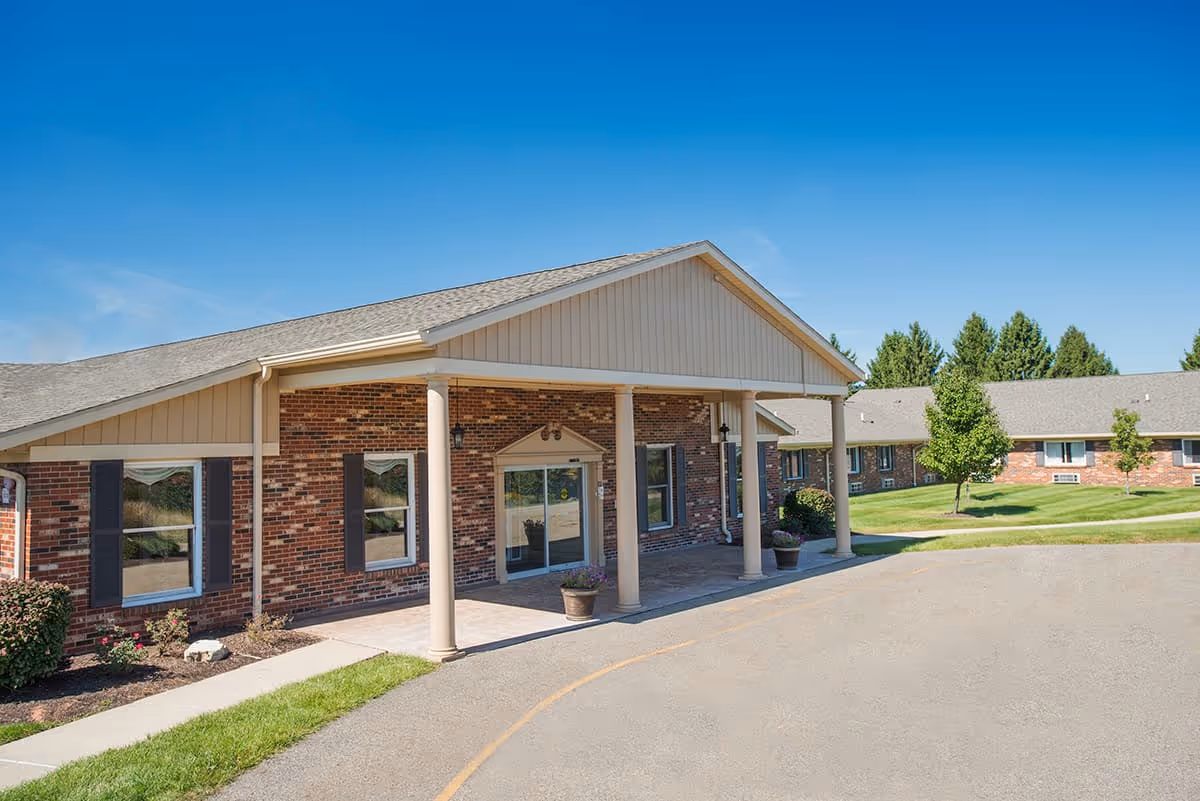 Front entrance of a single-story brick senior living facility with a covered portico, landscaping, and a clear blue sky.