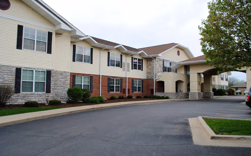 Exterior front facade and covered entrance of a two-story senior living building with brick and siding, landscaping, and a driveway.