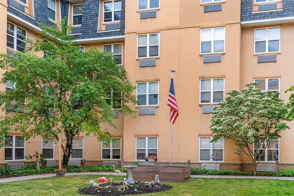 Outdoor courtyard area of a senior living facility with a green lawn, two trees, a flower bed with garden decorations, and an American flag on a flagpole. The courtyard is surrounded by a multi-story building with numerous windows.