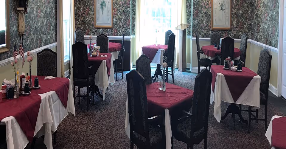 Dining room with multiple tables covered in white and burgundy tablecloths, each set with condiments and flowers in vases. The room has patterned wallpaper, carpeted floor, and large windows letting in natural light.