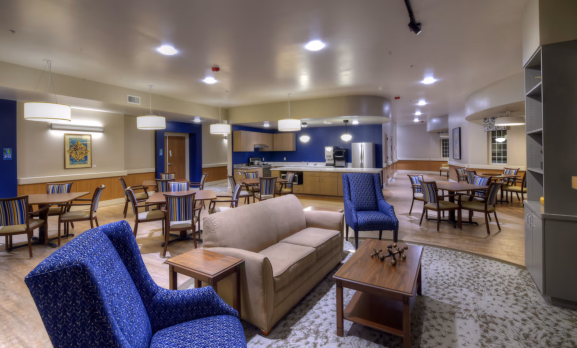 A spacious senior living community common area featuring a seating arrangement with a beige sofa, two blue patterned armchairs, and a wooden coffee table on a patterned rug. In the background, there are multiple dining tables with chairs, pendant lights hanging from the ceiling, and a kitchen area with wooden cabinets, a refrigerator, and a coffee machine.