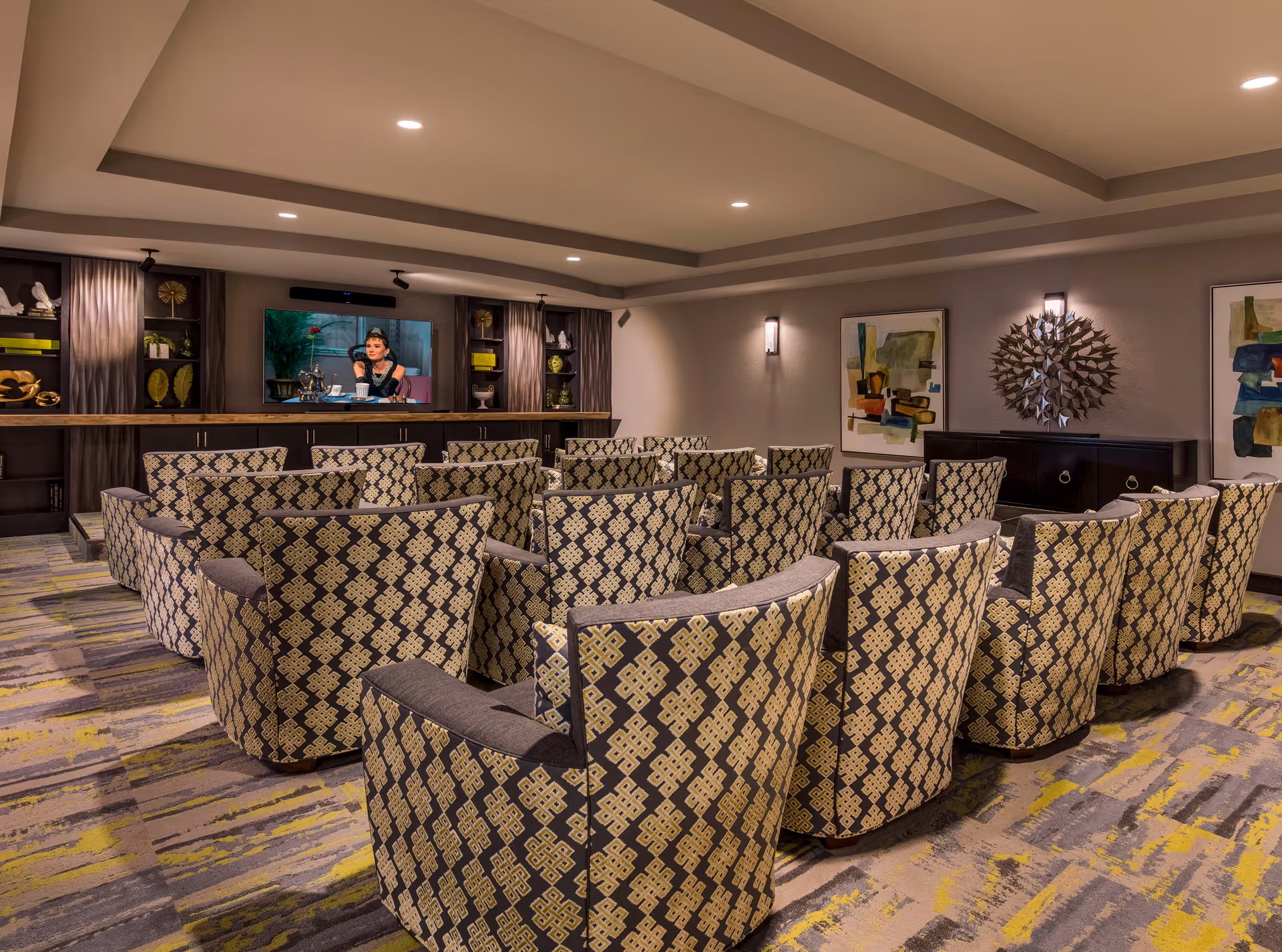 A cozy media room with multiple patterned armchairs arranged in rows facing a large flat-screen TV mounted on a wall with built-in shelves and cabinets. The room features modern ceiling lights, abstract artwork on the walls, and decorative items on the shelves and cabinets.