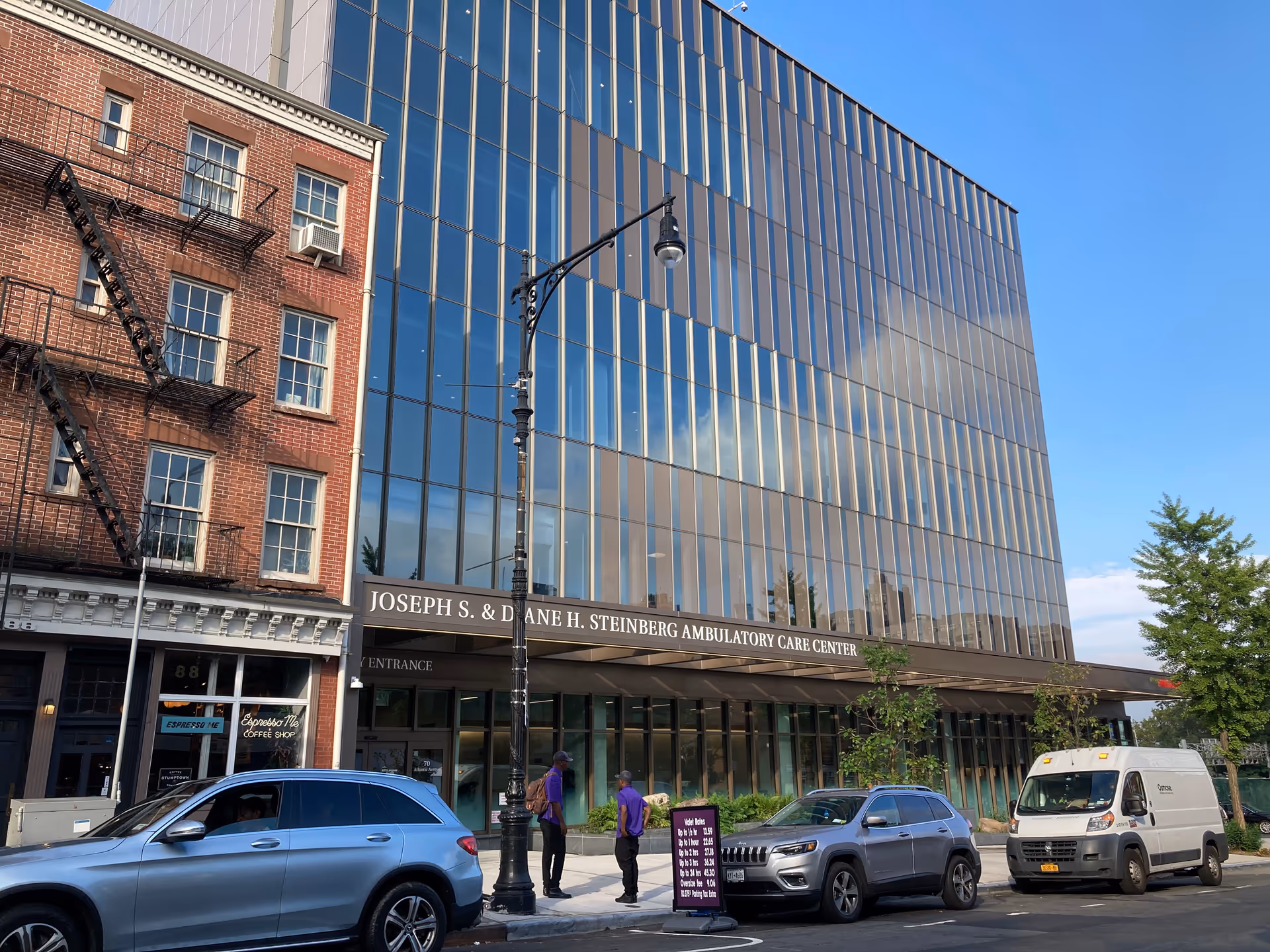 Glass-front ambulatory care center facade with adjacent brick building and parked cars.