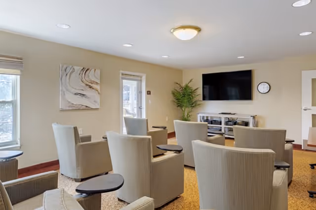 A small meeting or media room with several beige armchairs arranged in rows facing a wall-mounted flat screen TV. The room has beige walls, a carpeted floor, a potted plant in the corner, a wall clock, and a piece of abstract artwork. There are windows letting in natural light and a door leading outside.