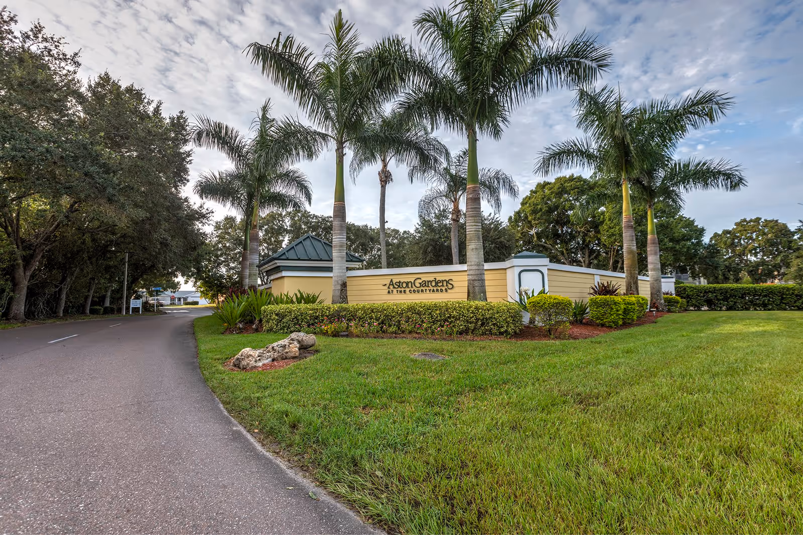 Entrance sign for Aston Gardens At The Courtyards surrounded by palm trees and well-maintained landscaping, with a paved road curving to the left and a partly cloudy sky overhead.