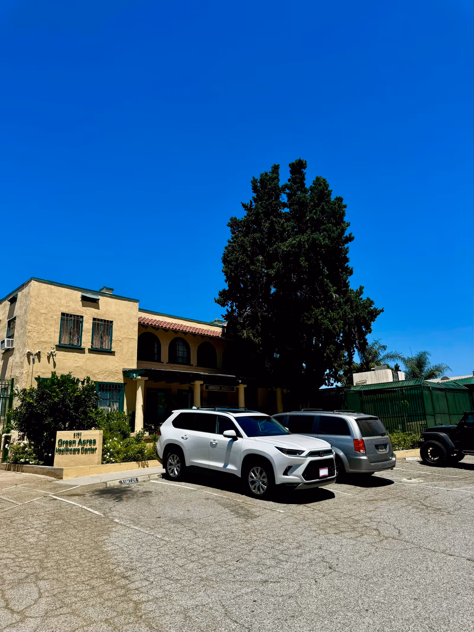 Beige senior living facility front with parked cars and a tall tree under a blue sky.