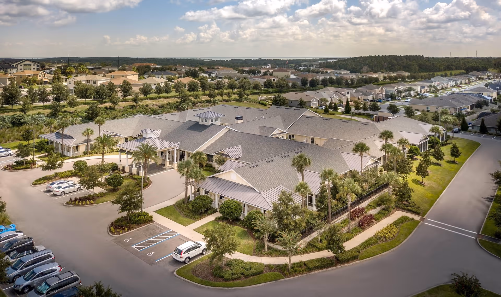 Aerial view of Benton House of Clermont senior living facility showing a large single-story building surrounded by landscaped greenery, palm trees, parking lots with cars, and nearby residential houses under a partly cloudy sky.