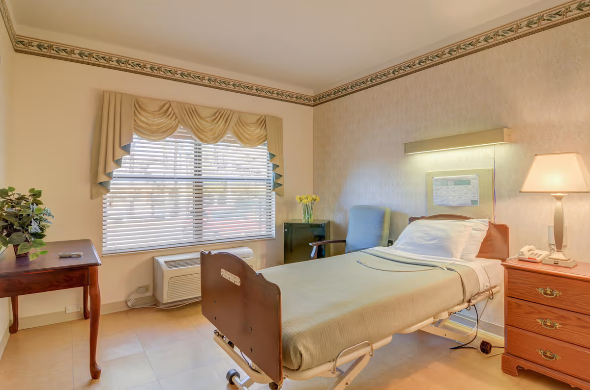 Single furnished bedroom with a hospital-style adjustable bed, window blinds, lamp, and dresser.