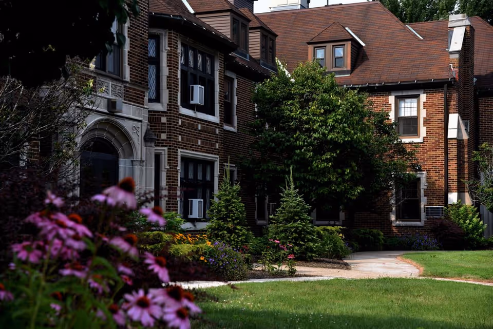 Brick building with arched entrance, curved path, and colorful flower beds in a landscaped yard.
