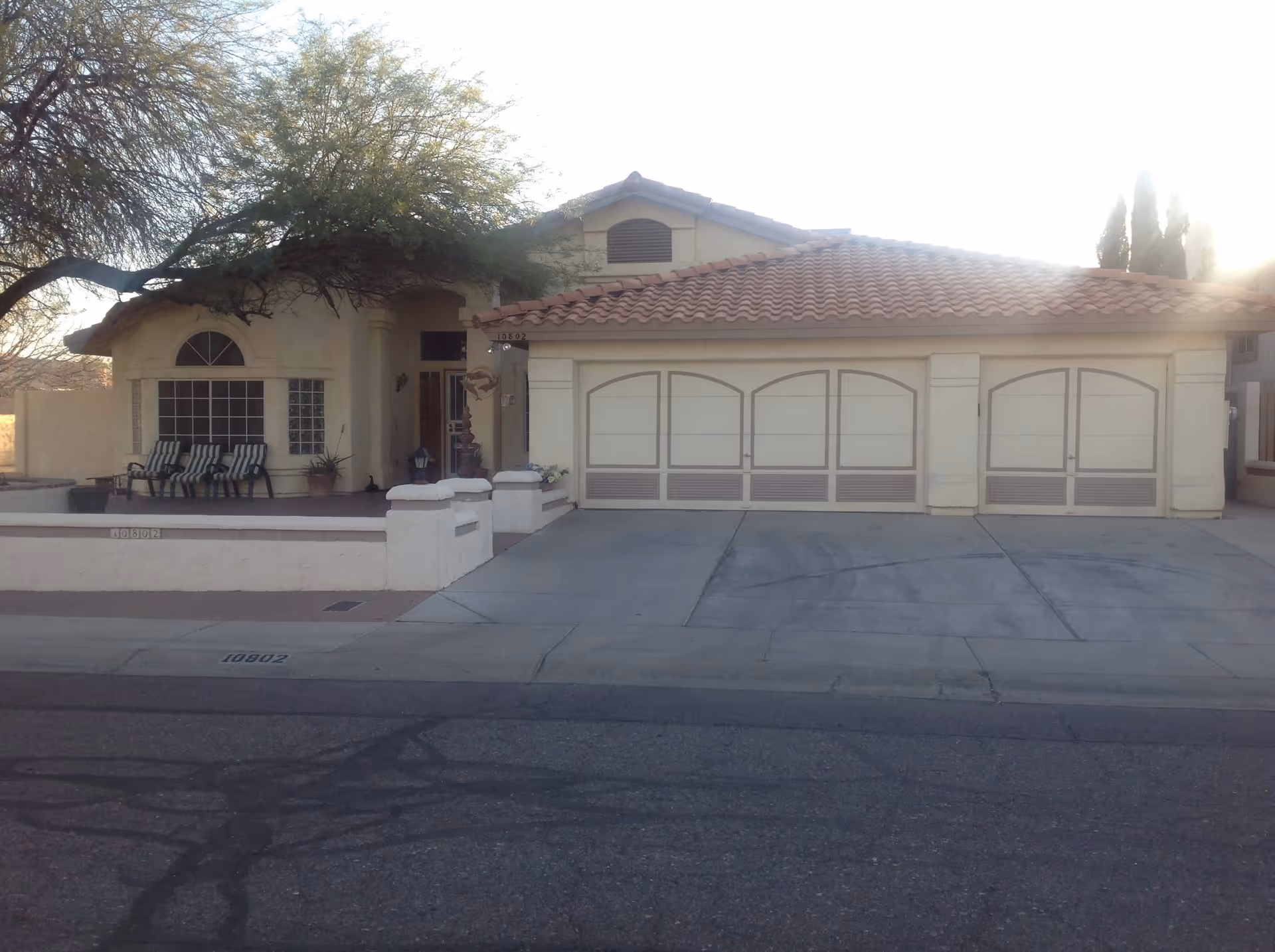 Single-story stucco house with a tiled roof, three-car garage, front porch with chairs and a low wall, partially shaded by a tree.