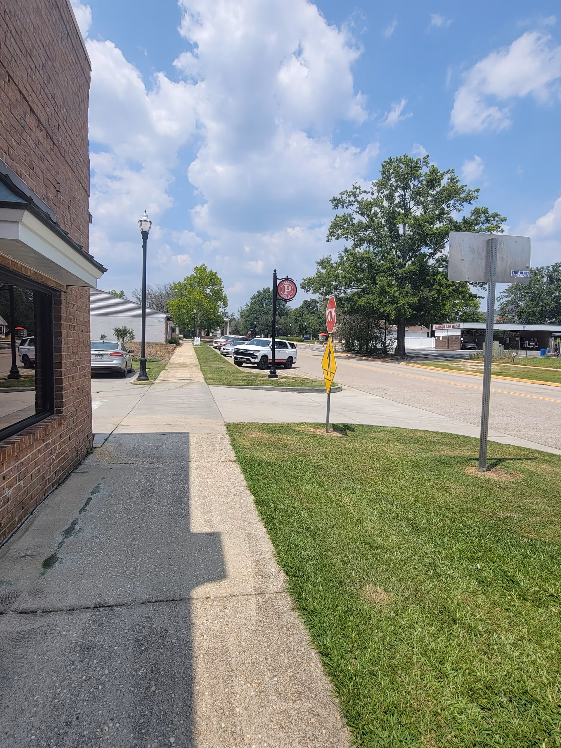 Sidewalk runs beside a brick building with street lamps, cars, and green lawns under sunny sky.