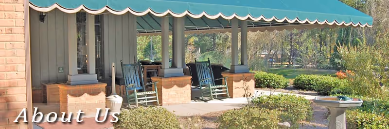 Covered outdoor patio area with rocking chairs and brick pillars at Shadow Oaks Assisted Living, surrounded by bushes and greenery.