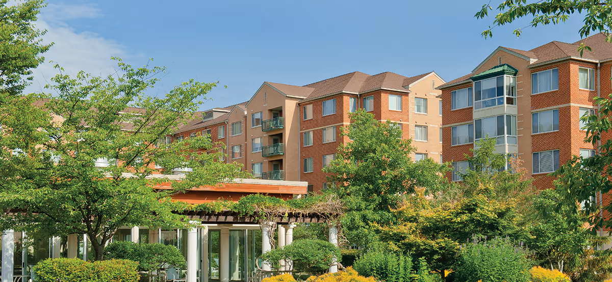 Exterior view of a multi-story brick senior living building with landscaped gardens and a pergola-covered entrance.