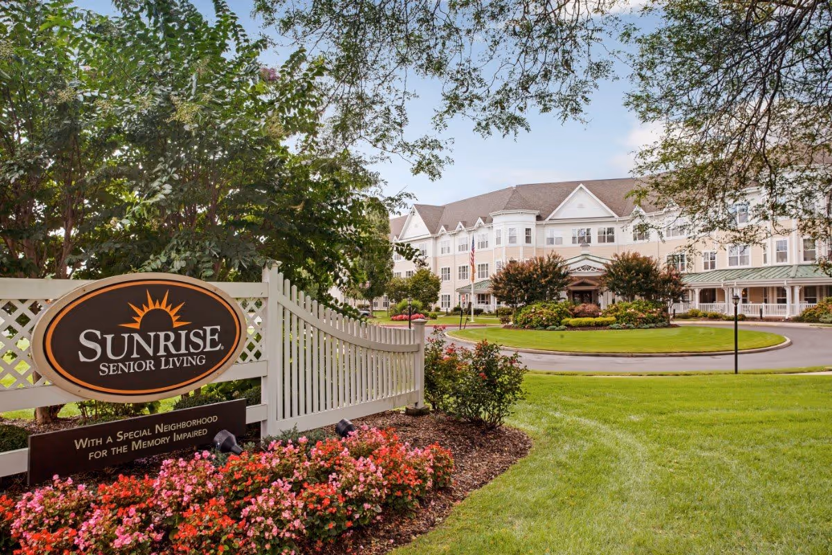 Front entrance of Sunrise Senior Living with a landscaped lawn, white fence and sign in the foreground and the facility building in the background.