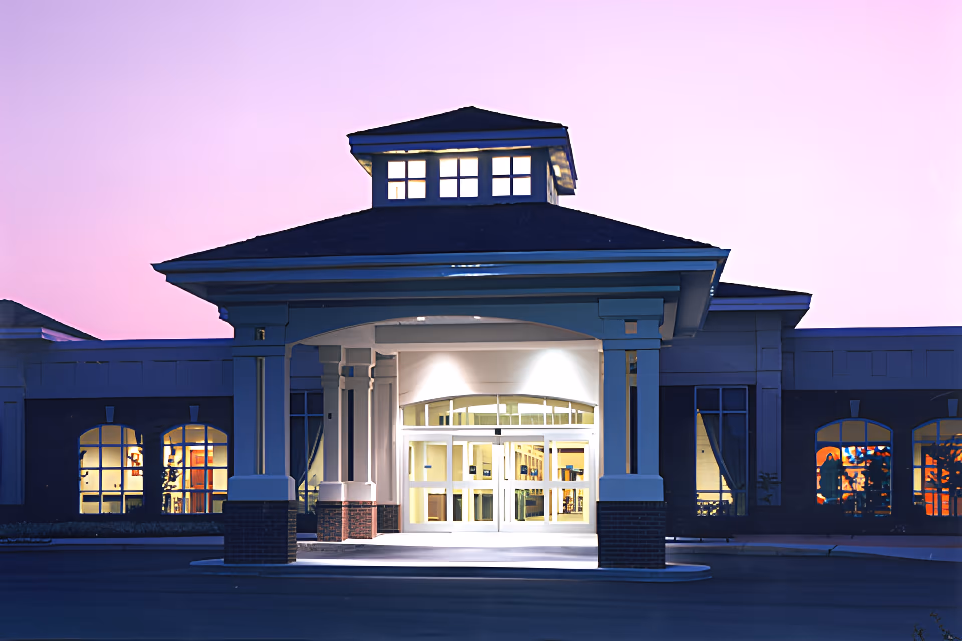 Front entrance of a building with a covered drop-off area supported by columns, illuminated at dusk with a purple sky in the background. The building has large windows with warm interior lighting visible inside.
