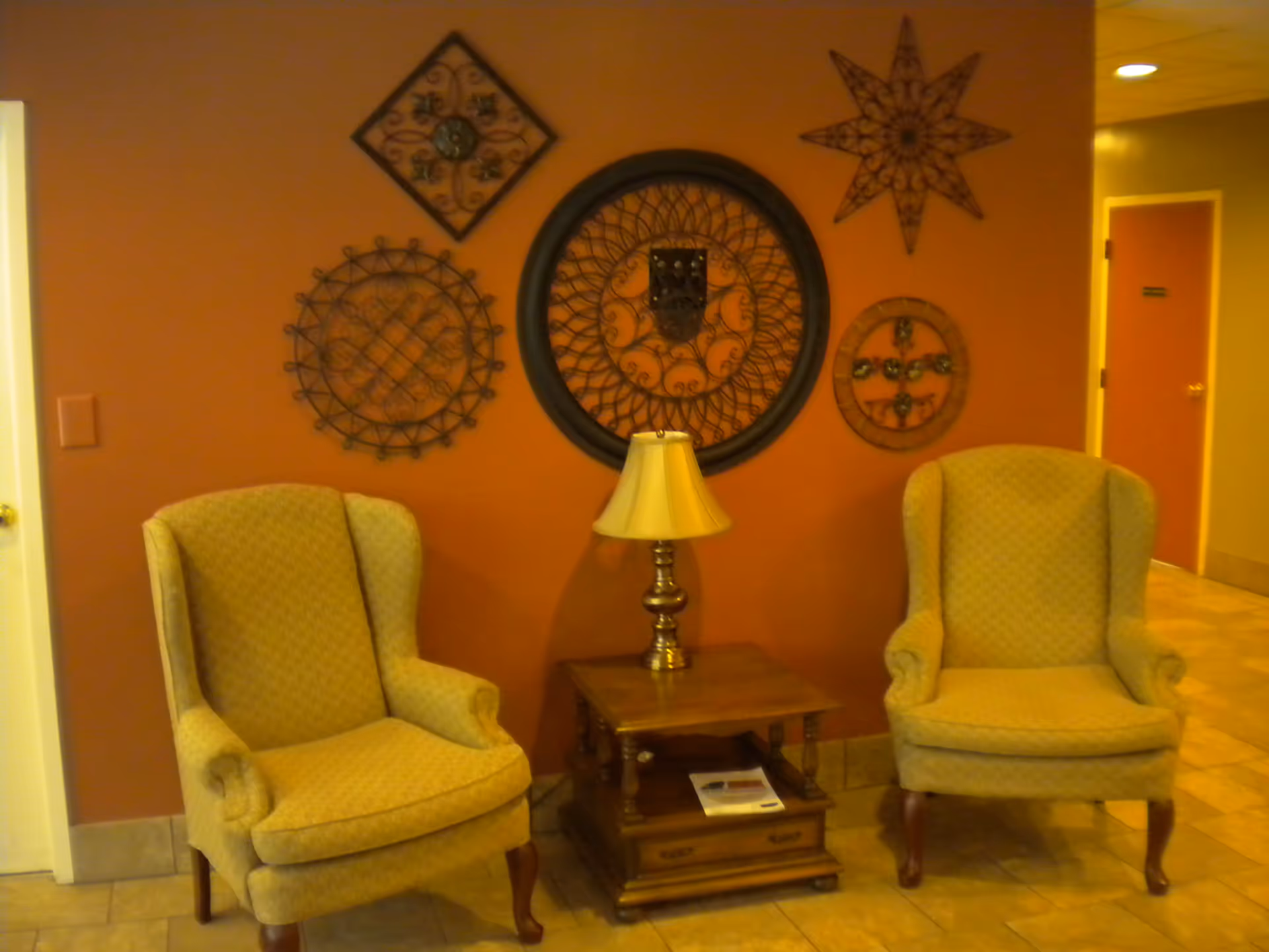 Two upholstered armchairs flank a wooden side table with a lamp in front of a wall decorated with circular metal wall art.