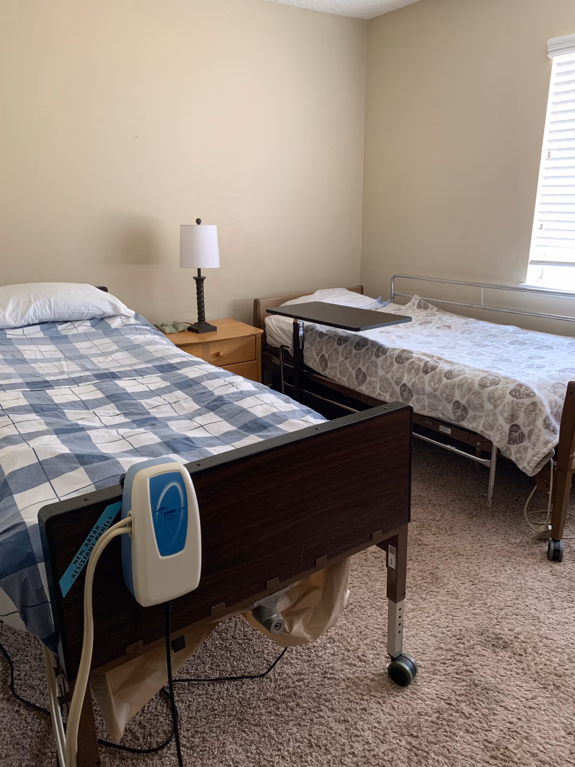 Two hospital-style beds with a nightstand, lamp, and window in a shared bedroom.