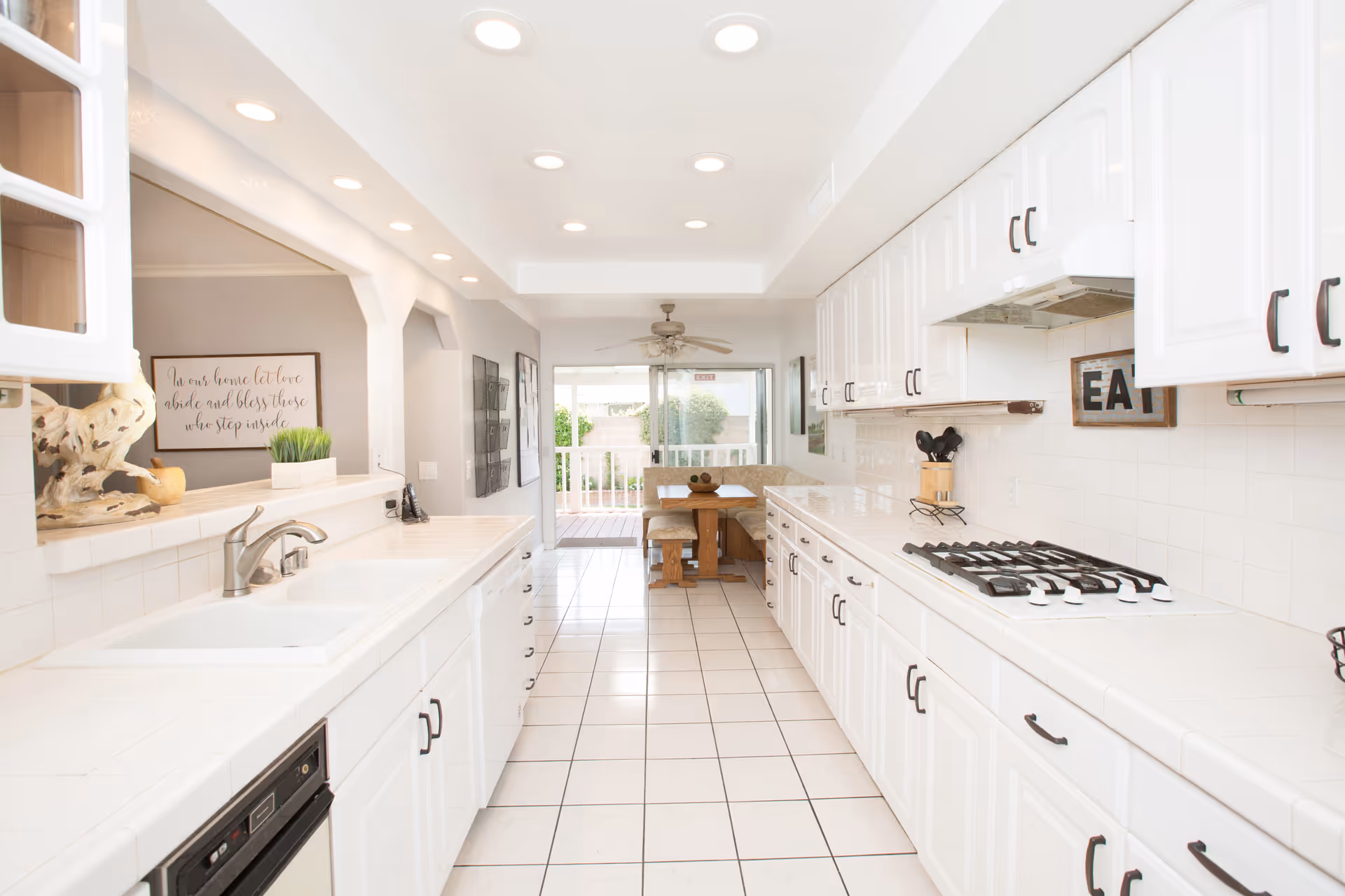 Bright and clean kitchen with white cabinets and tiled countertops. The kitchen features a gas stove, a sink, and a dishwasher. At the far end, there is a cozy dining nook with a wooden table, bench seating, and a ceiling fan. Large sliding glass doors lead to an outdoor area with greenery visible.