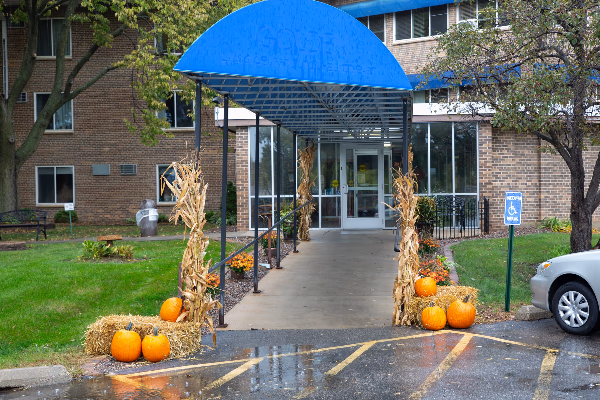 Entrance to a brick building with a blue awning over a concrete walkway. The walkway is decorated with hay bales, pumpkins, and dried corn stalks, suggesting a fall or Halloween theme. There is a handicapped parking sign and a silver car parked nearby. Trees and green grass surround the entrance area.
