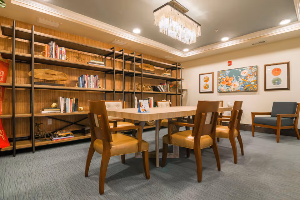 A cozy interior room with a rectangular table surrounded by six wooden chairs with tan cushions. Behind the table is a large shelving unit filled with books and decorative items. The walls are adorned with three framed floral artworks. The room is softly lit by a modern chandelier hanging from the ceiling.