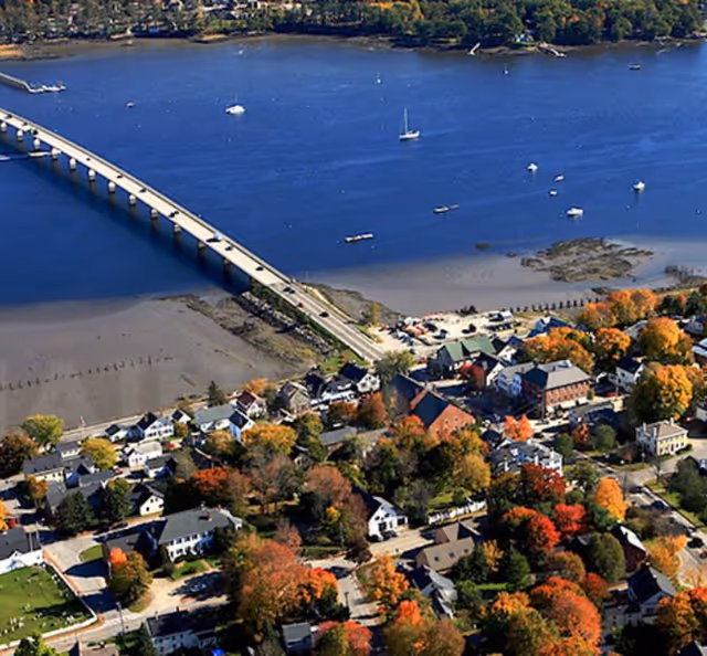 Aerial view of a coastal town with a long bridge, harbor, and autumn trees.