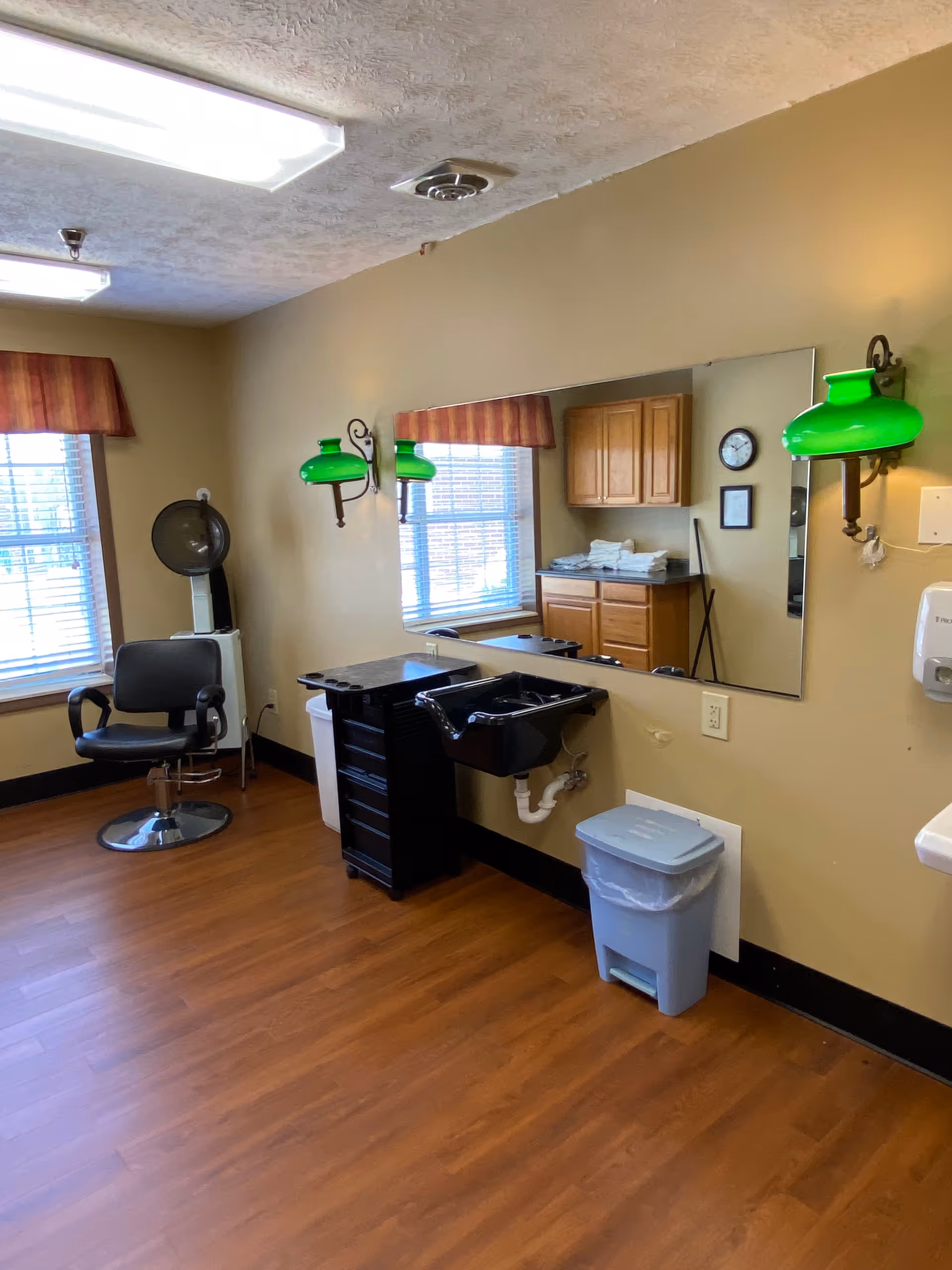 Indoor senior living facility salon with chair, mirrors, sinks, cabinets, and green lamps.