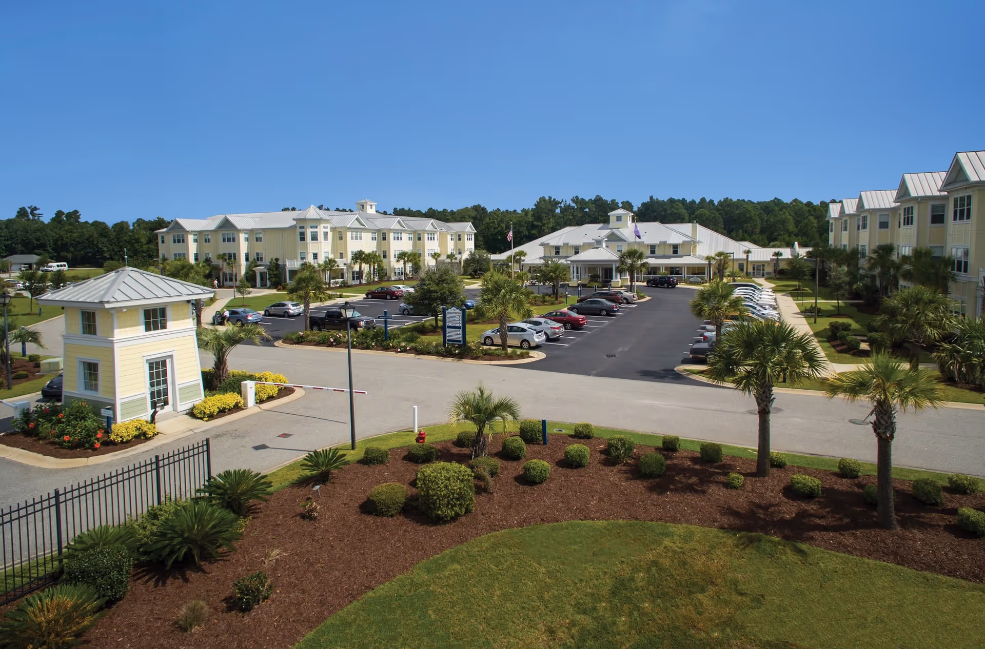 View of a senior living facility with multiple yellow buildings, a parking lot with cars, landscaped gardens with palm trees and shrubs, and a clear blue sky.