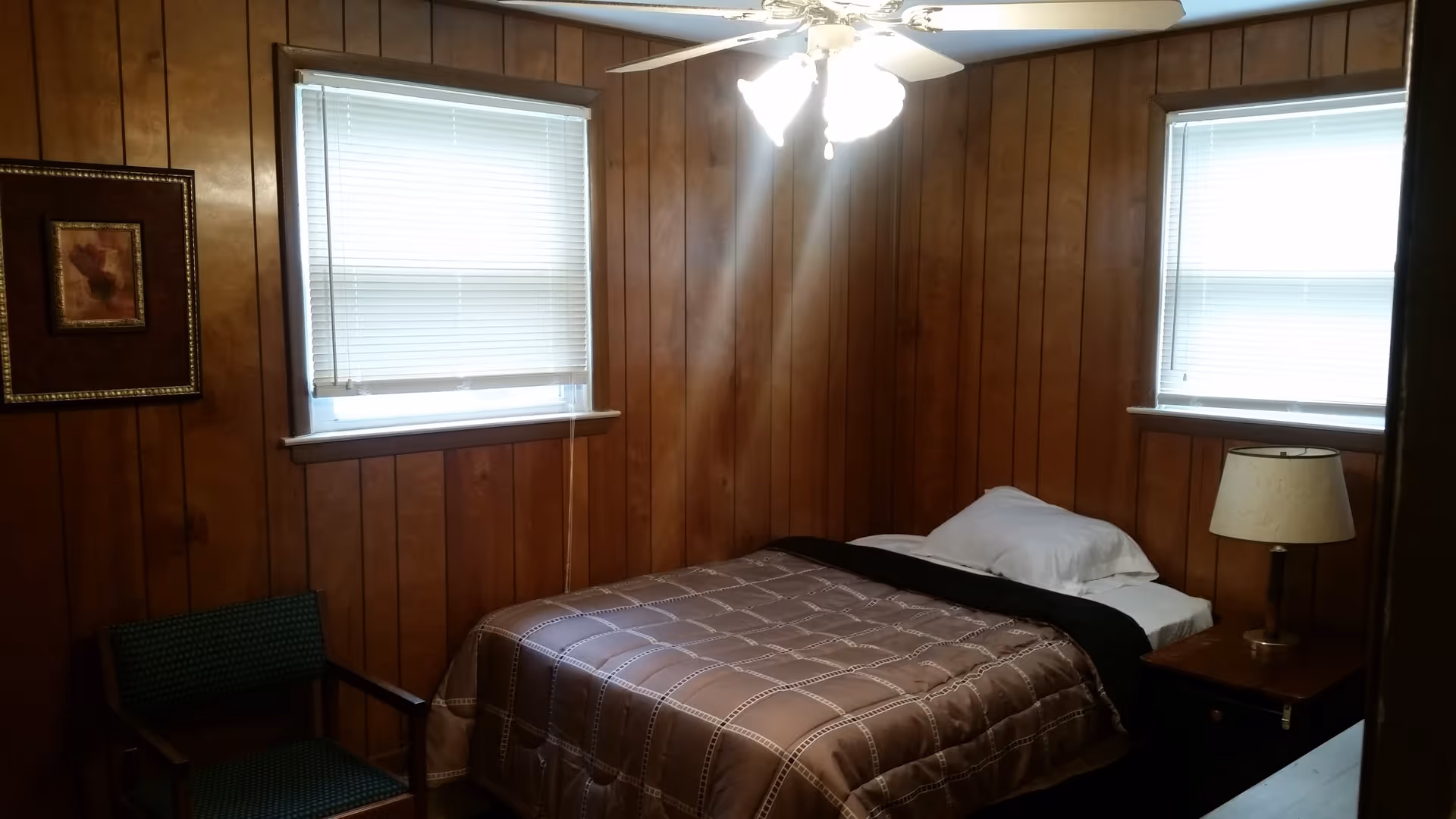 Small bedroom with wood-paneled walls, a single bed with a nightstand and lamp, a ceiling fan, two windows, and a chair.