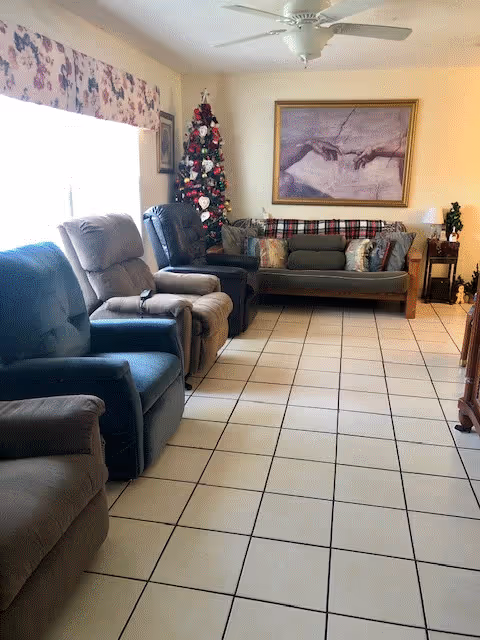 A cozy living room with several recliner chairs lined up along the left wall and a couch at the far end. A decorated Christmas tree stands in the corner near the couch. Above the couch is a framed artwork depicting two hands reaching towards each other. The room has tiled flooring, a ceiling fan, and a window with floral curtains on the left side.