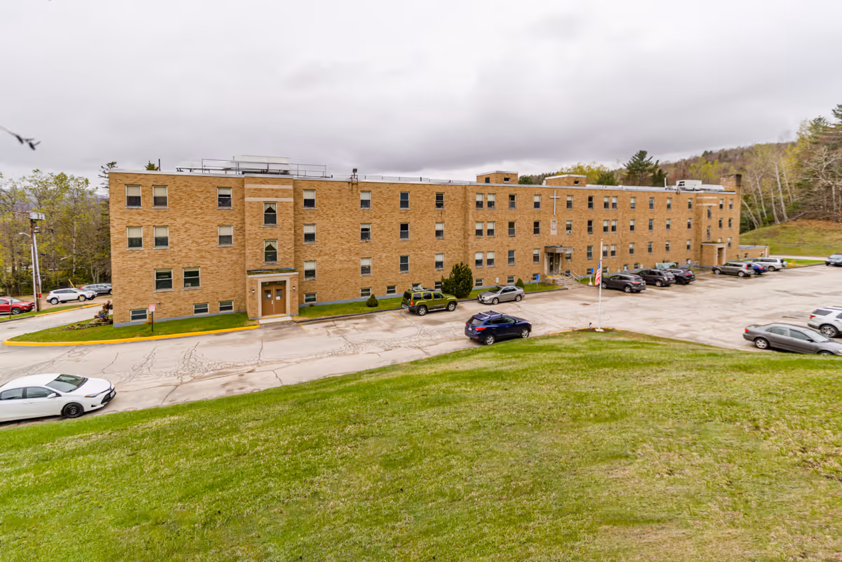 Exterior view of a three-story brick building with multiple windows and two main entrances. Several cars are parked in front of the building on a paved parking lot. There is a grassy hill in the foreground and trees in the background under a cloudy sky.