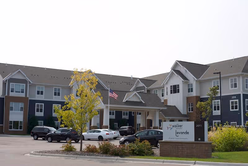 Exterior view of The Levande by Vivie senior living facility showing a large three-story building with multiple windows, a covered entrance with an American flag, several parked cars, and a sign in front displaying the facility's name and address.
