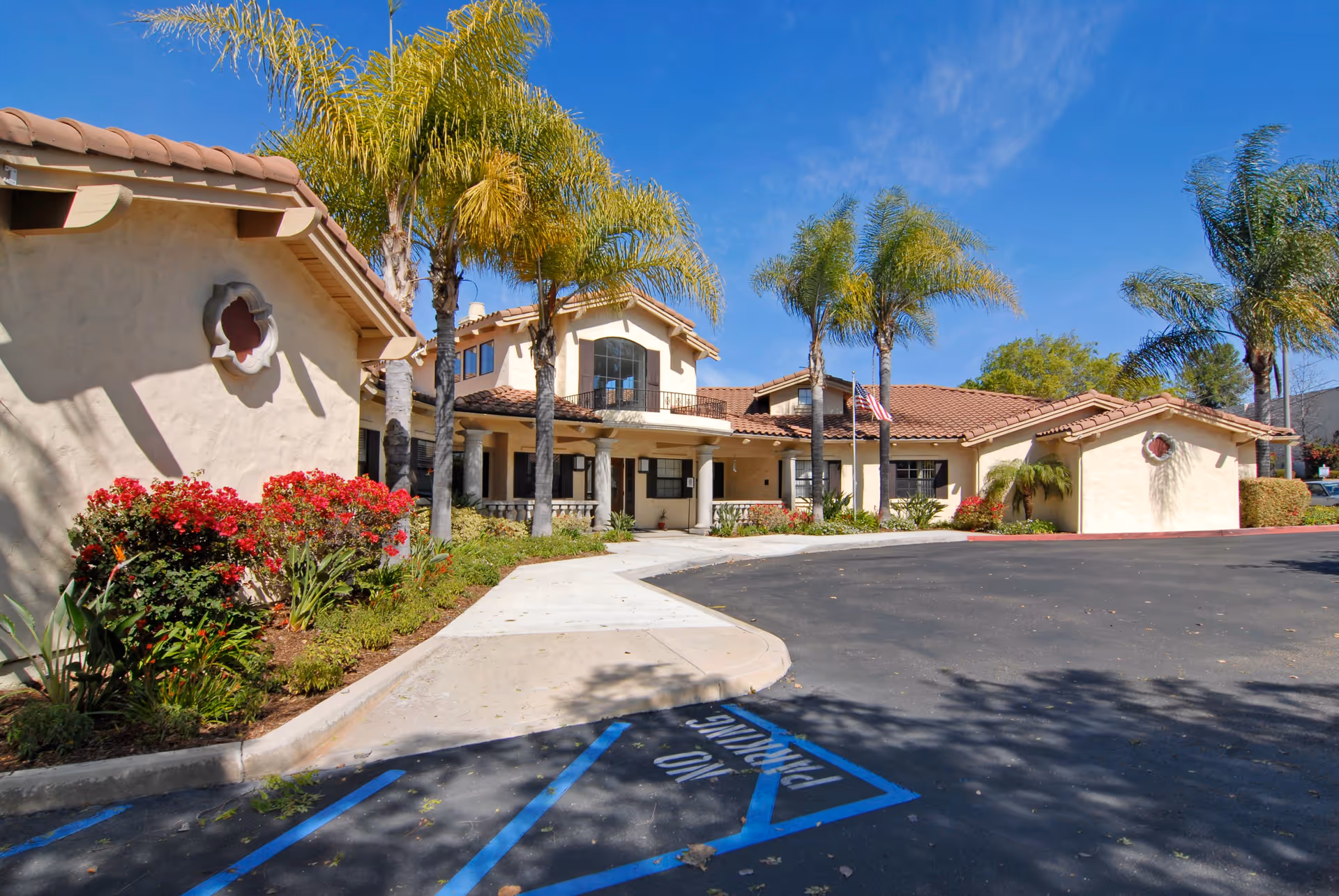 Exterior view of Grossmont Gardens Memory Care facility showing a beige stucco building with a red tile roof, palm trees, and landscaped bushes with red flowers under a clear blue sky. A curved driveway and a sidewalk lead to the entrance.