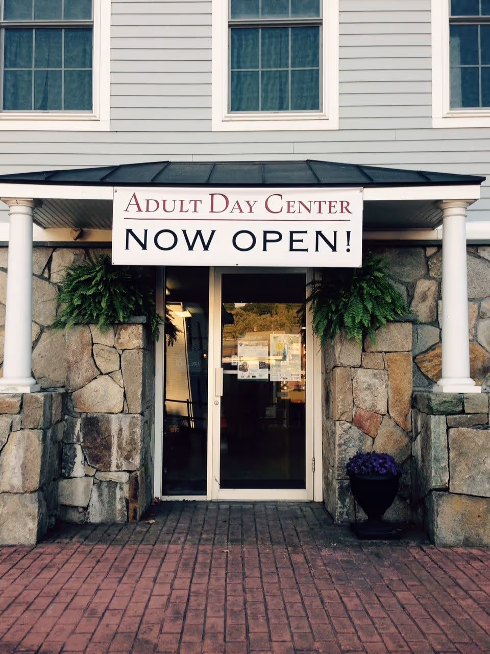 Entrance to a stone-faced building with a banner reading 'Adult Day Center NOW OPEN!' above glass double doors.
