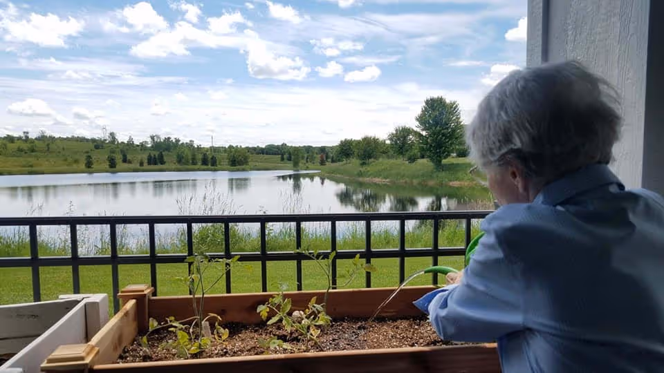 An elderly person on a balcony tending a planter with a lake view.