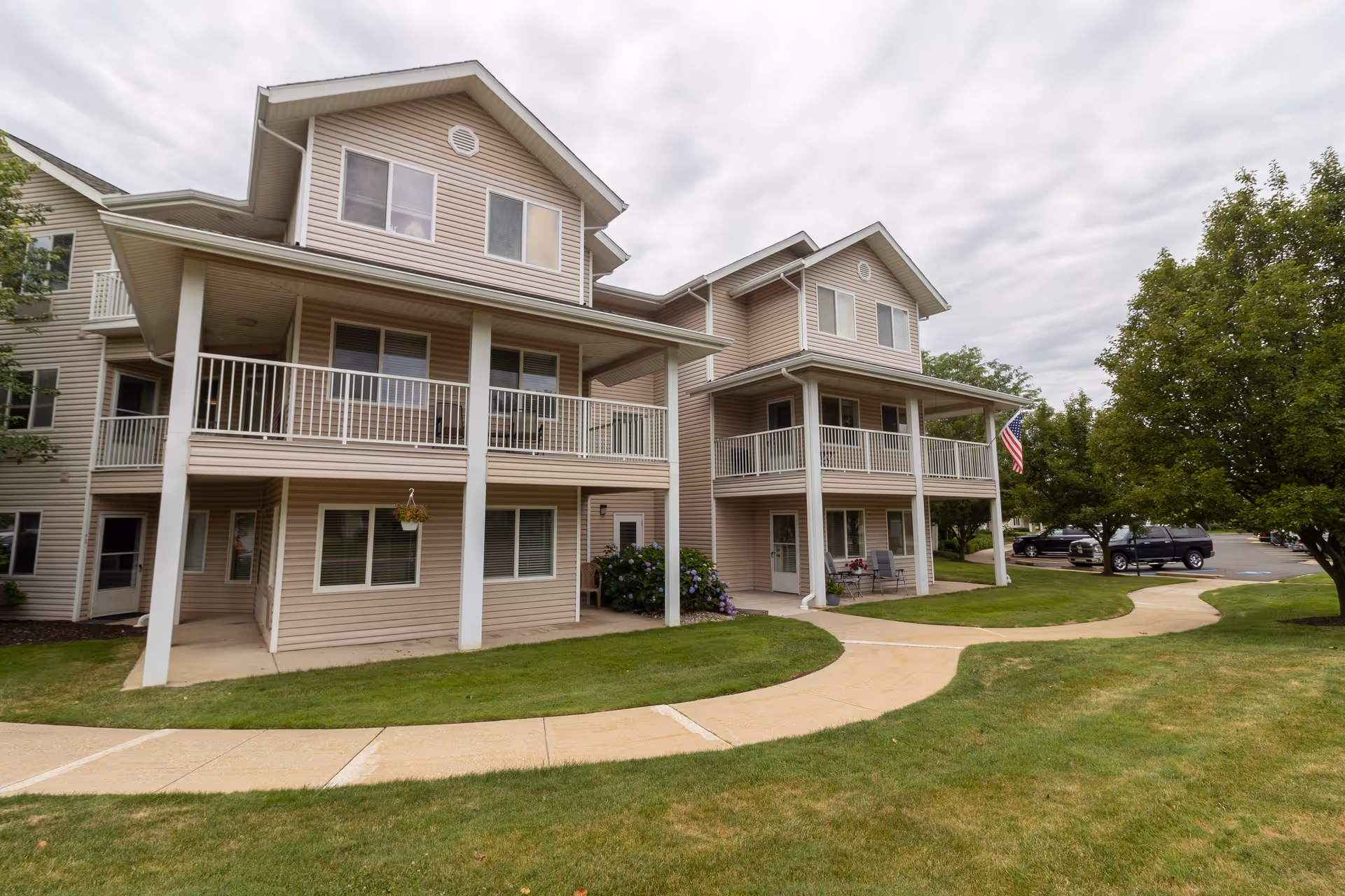 Exterior view of a two-story residential building with beige siding and white railings on the balconies. There is a curved concrete walkway in front of the building, green grass, and trees. Several parked cars are visible in the background under a cloudy sky.