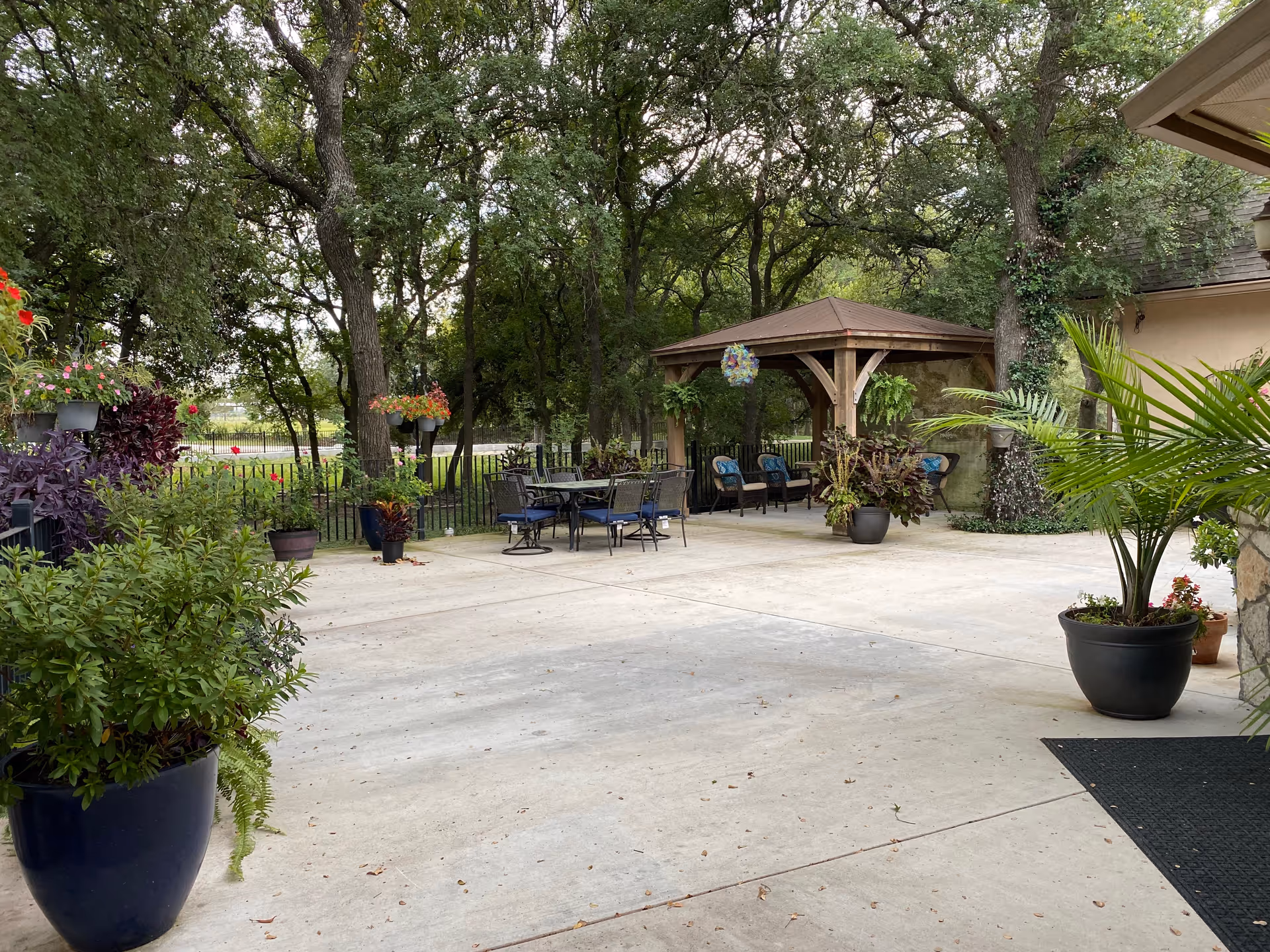 Outdoor patio area with several potted plants and flowers, a table with chairs, and a wooden gazebo with cushioned seating. Trees and greenery surround the space, creating a peaceful and shaded environment.