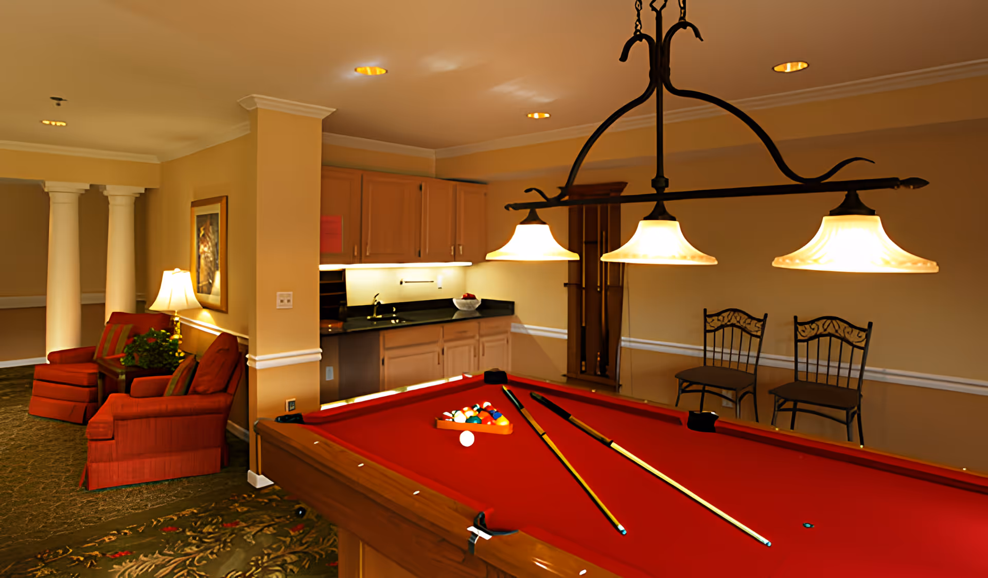 Interior view of a recreational room featuring a red pool table with billiard balls and cues, two hanging pendant lights above the table, a small kitchenette with wooden cabinets and a black countertop in the background, two decorative chairs against the wall, and a seating area with red upholstered armchairs and a lamp near white columns.
