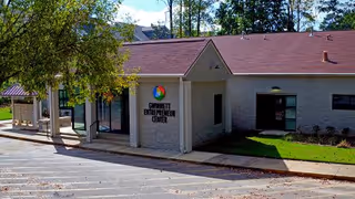 Exterior view of a single-story building with a sign, parking lot, trees, and lawn.