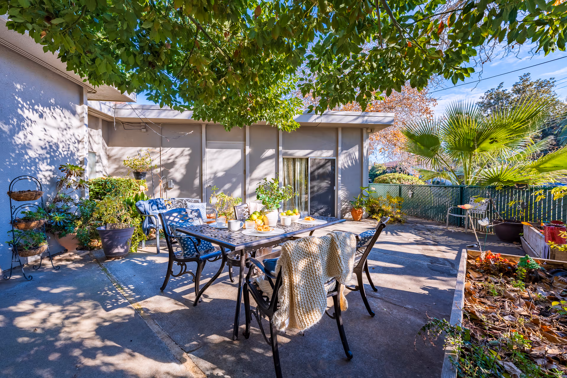 Outdoor patio area with a metal table and chairs, some chairs draped with knitted blankets. The table is set with cups, plates, and a bowl of fruit. Surrounding the patio are various potted plants and greenery, with a large tree providing shade overhead. A sliding glass door leads into the building, and a fence encloses the area with more plants and trees visible beyond.