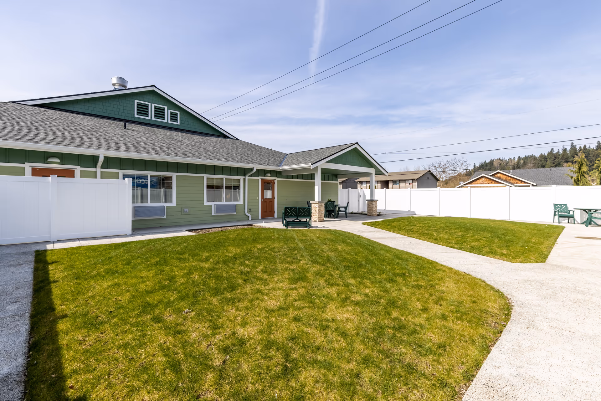 Outdoor view of a senior living facility named Memory Haven featuring a green lawn, concrete pathways, white fencing, and a green building with a covered porch area with benches and chairs under a clear sky.