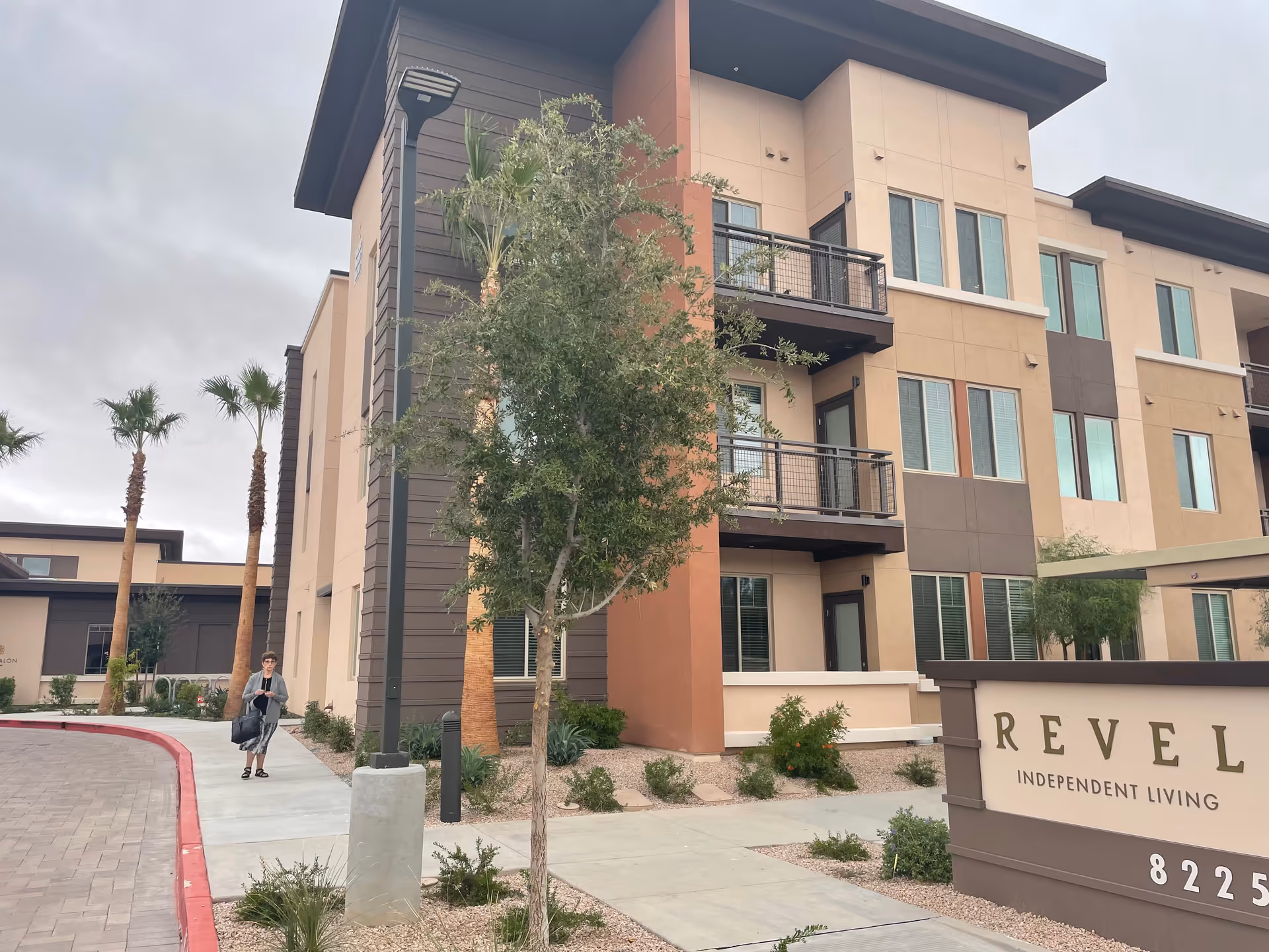 Three-story modern senior living building with a sidewalk, trees, and a Revel Independent Living sign.