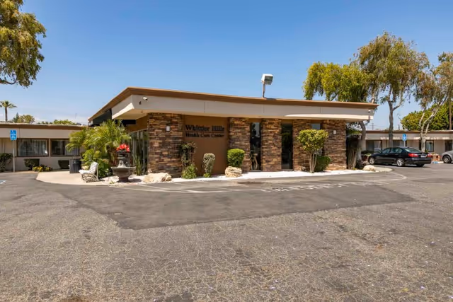 Front exterior of the Whittier Hills Health Care Center building with a parking lot and landscaping under a clear blue sky.