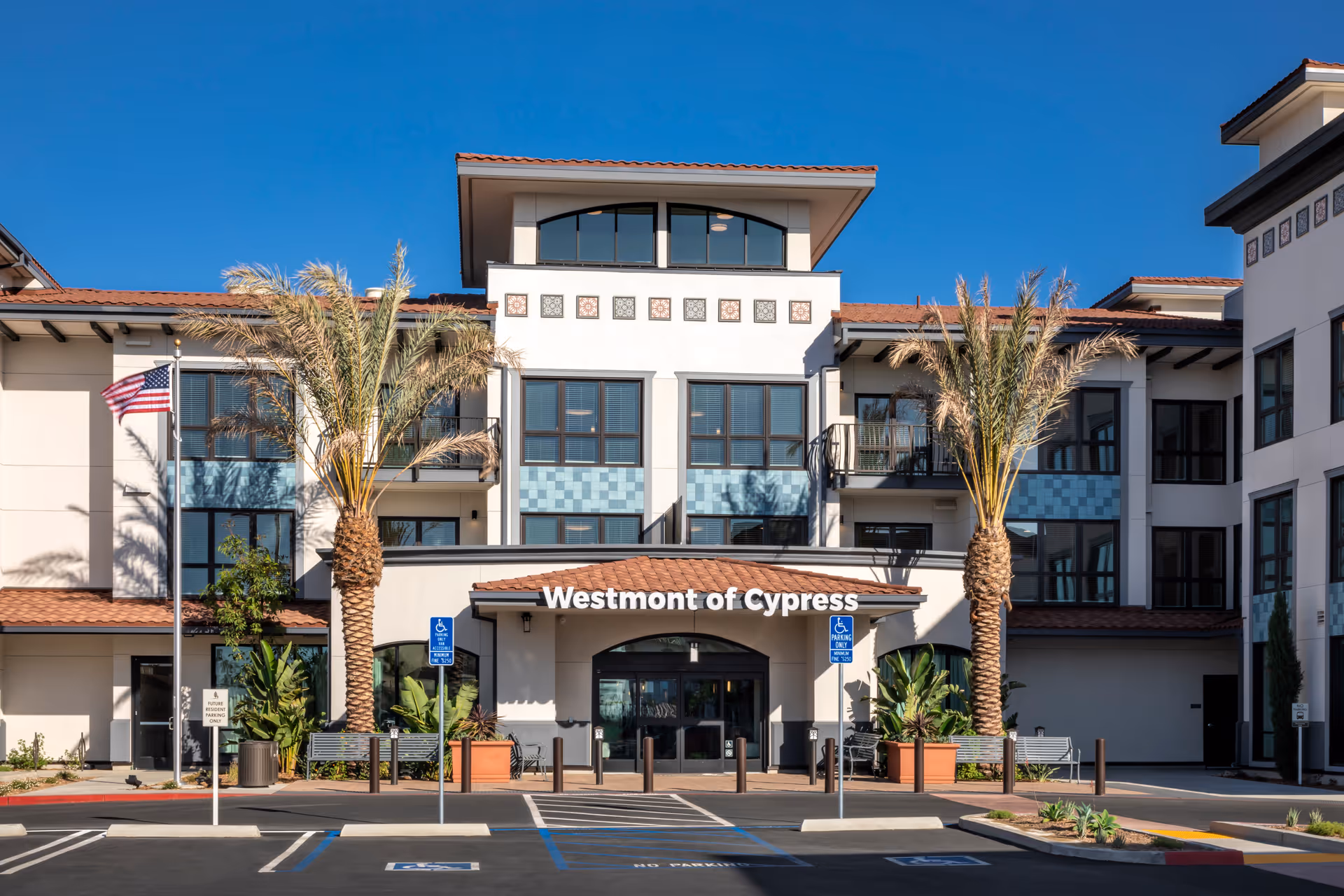 Front exterior view of Westmont of Cypress senior living facility with palm trees, handicap parking spaces, and an American flag under a clear blue sky.