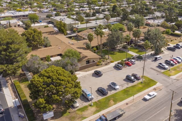 Aerial view of Pueblo Springs Rehabilitation Center showing multiple single-story buildings with brown roofs surrounded by trees and parking lots with several parked cars. The facility is located in a suburban area with residential houses and streets visible in the background.