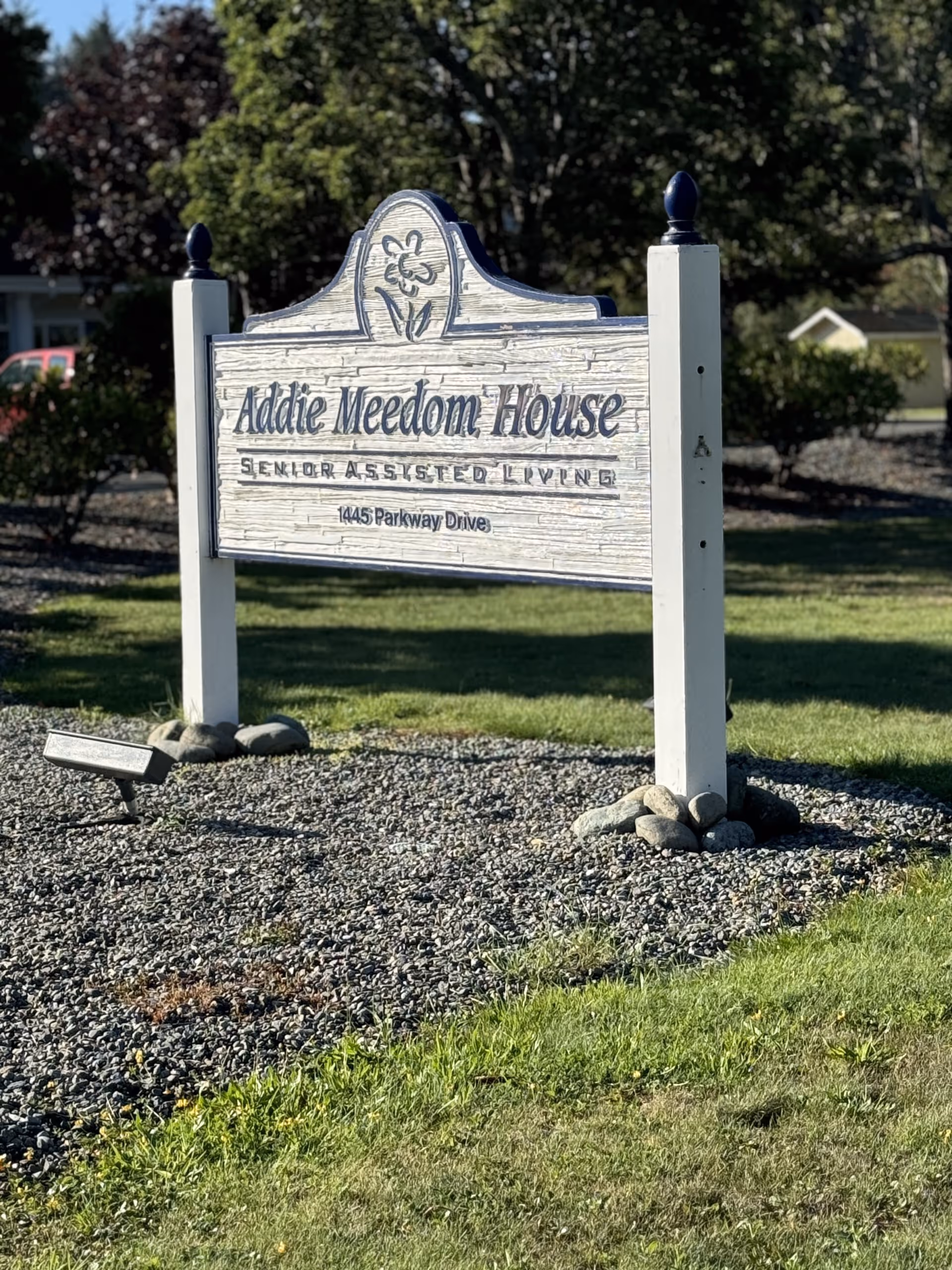 A wooden sign reading Addie Meedom House stands on a gravel lot with landscaping.