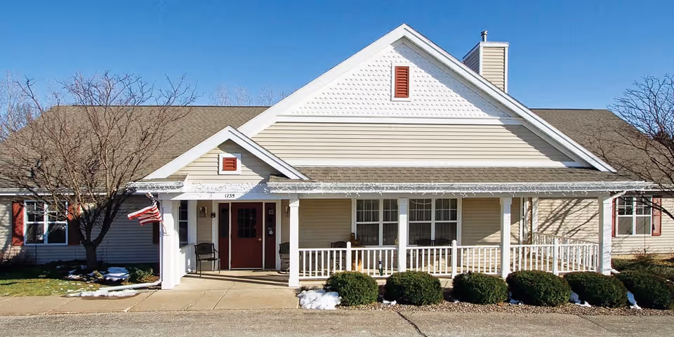 Front exterior view of a single-story senior living facility building with beige siding, a covered porch with white railings, two chairs near the entrance, an American flag, and some bushes and leafless trees in front under a clear blue sky.